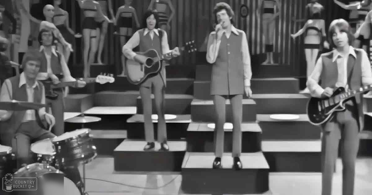 A mid-20th-century musical performance with three guitarists, a drummer, and mannequins in the background, set on an indoor stage with geometric patterns.