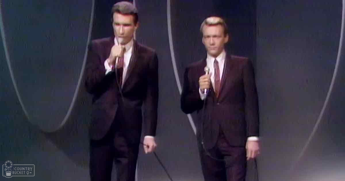 Two adult male singers dressed in formal dark wool suits with classic fits, holding microphones on an indoor stage at night.