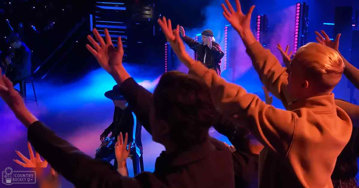 Young adults interacting on stage with performers at an evening concert; audience members in dark jackets, performers wearing black jackets and hats under blue-purple lighting.