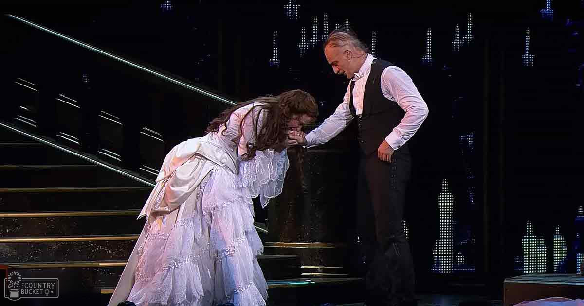 A theatrical scene in an urban opera house at night, with two actors dressed in historical costumes against a backdrop of illuminated city buildings.