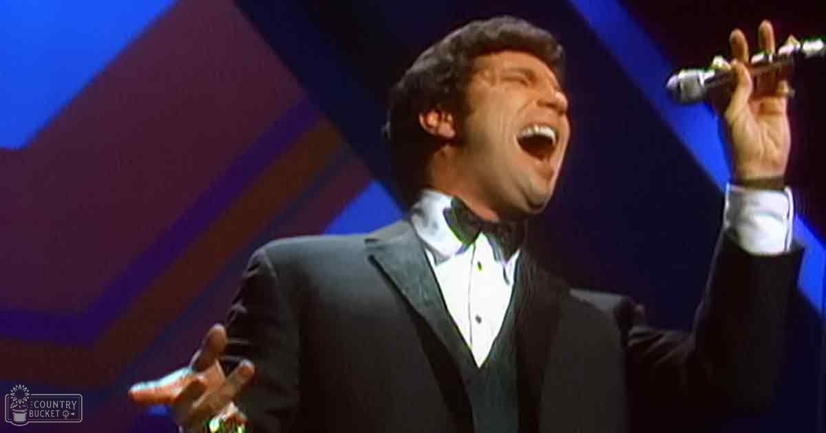 Adult male singer in a dark suit with white shirt, black bow tie, holding a microphone above his head on stage with geometric blue and maroon backdrop.