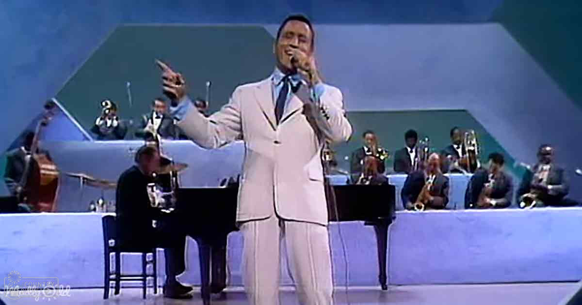 A male adult singer in a well-fitted mid-20th century light-colored suit with a tie and shirt, performing on stage at an orchestra setup.