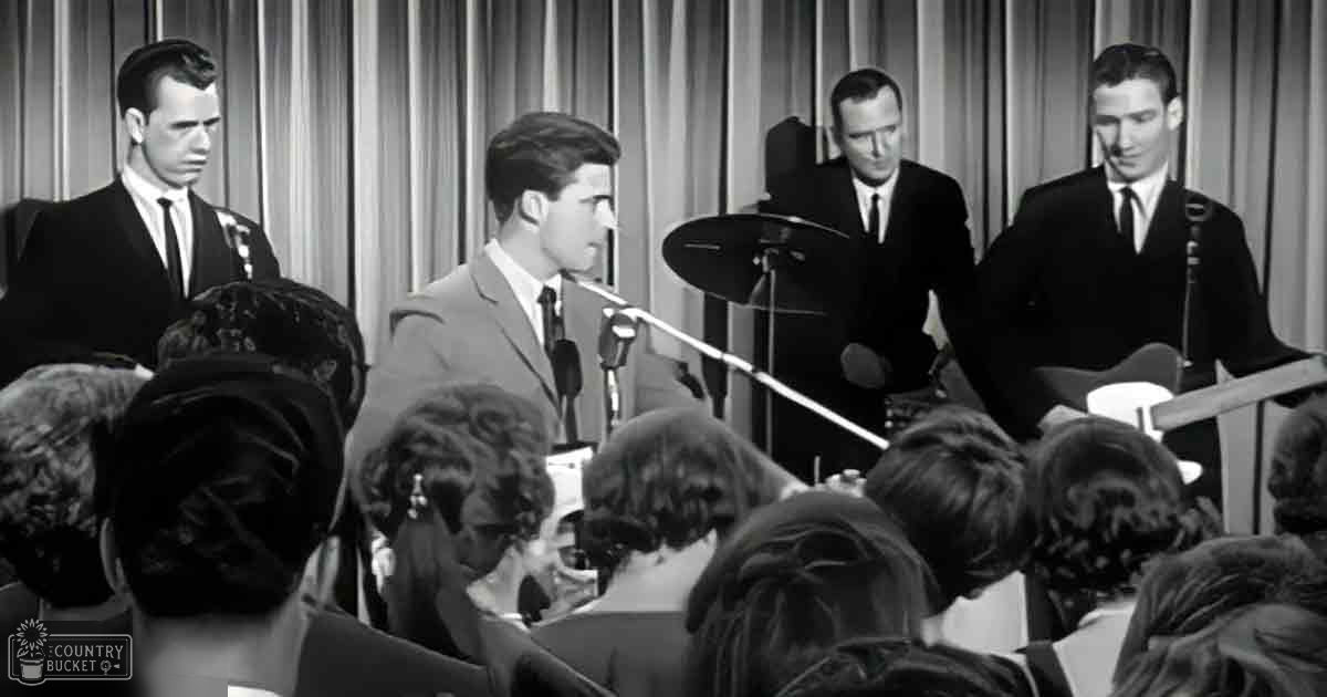 A young adult male singer performs on stage with musicians behind him; the audience appears diverse, dressed in various styles from an earlier era.