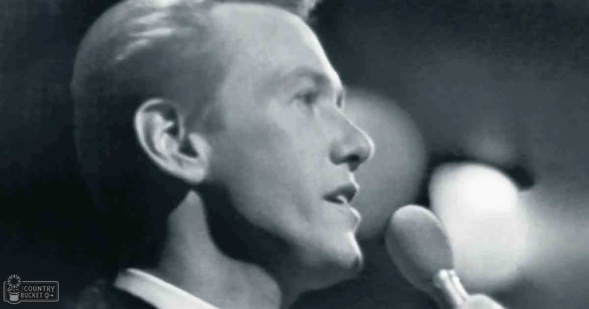 A middle-aged man speaks into a microphone at what appears to be an indoor concert or public speech venue.