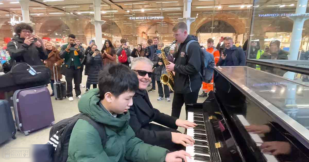 A young adult wearing a green puffer jacket plays piano at an indoor public space with glass walls, retail signs, and various individuals moving around.