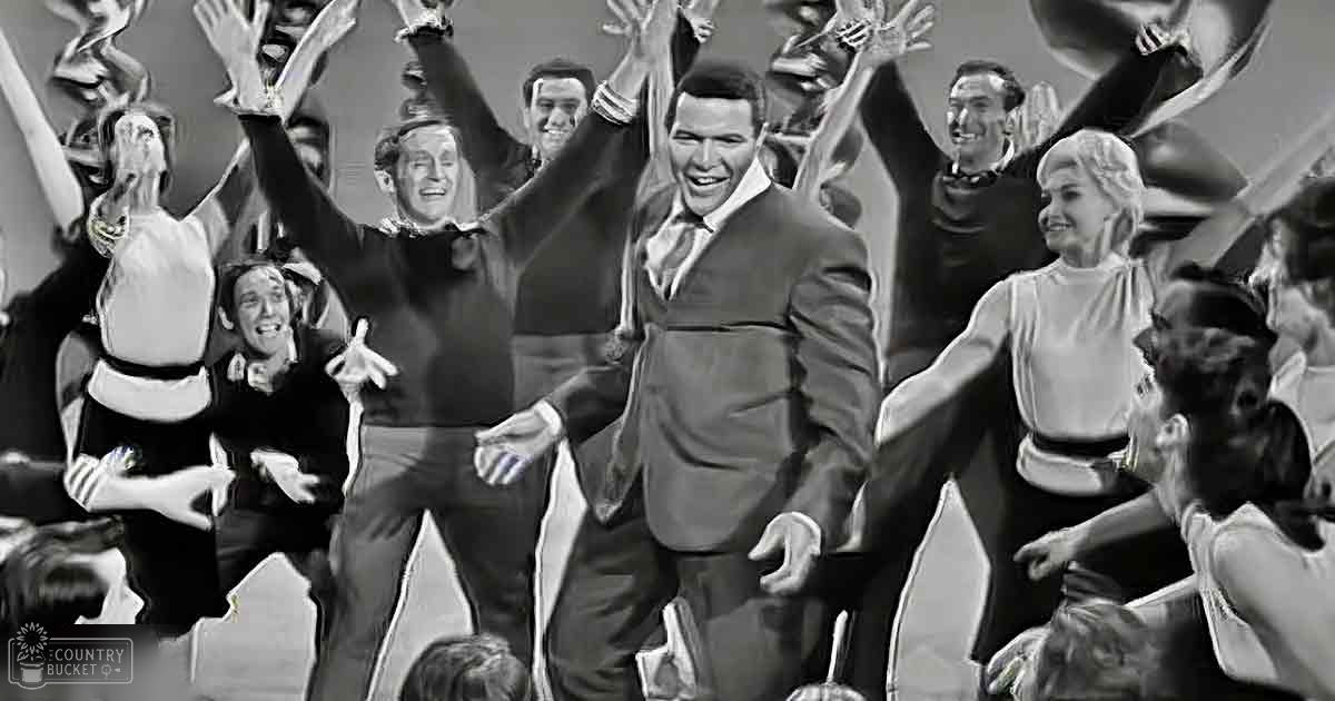 An adult male in a dark suit stands at center, surrounded by young adults and an older woman indoors with stage-like backdrop.