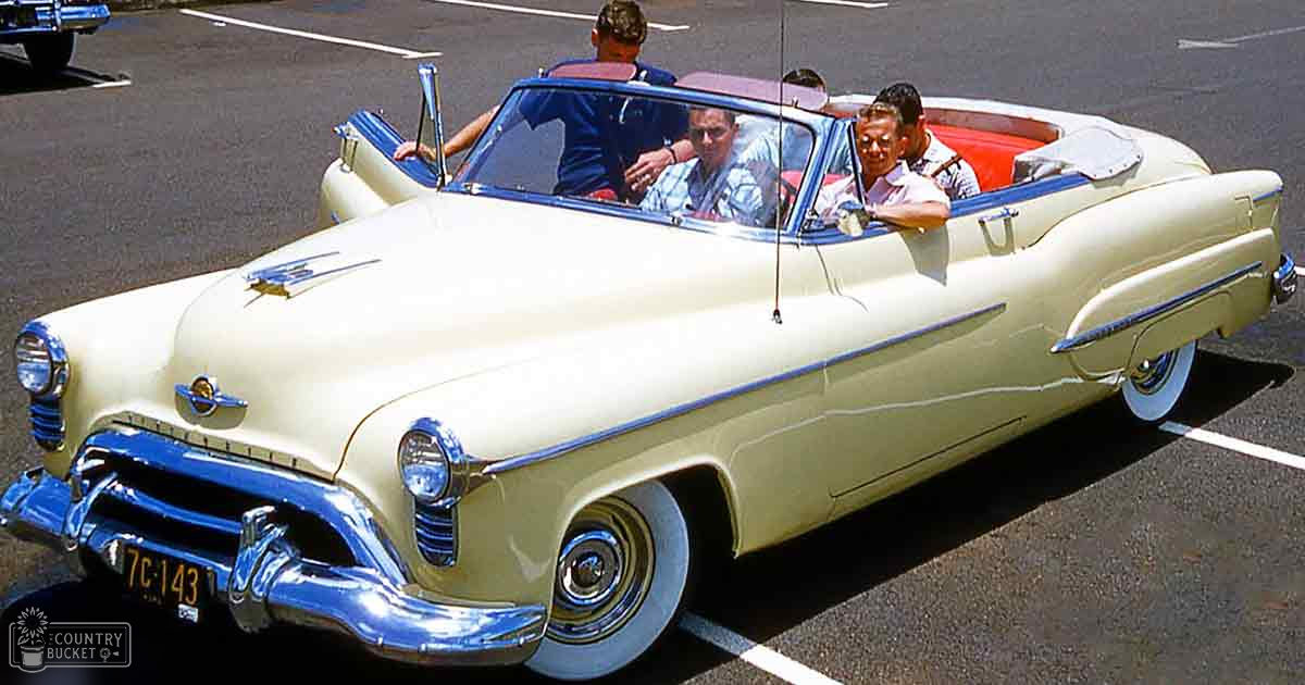A vintage convertible car with cream body and blue trim parked outdoors, surrounded by three adult males wearing casual attire.