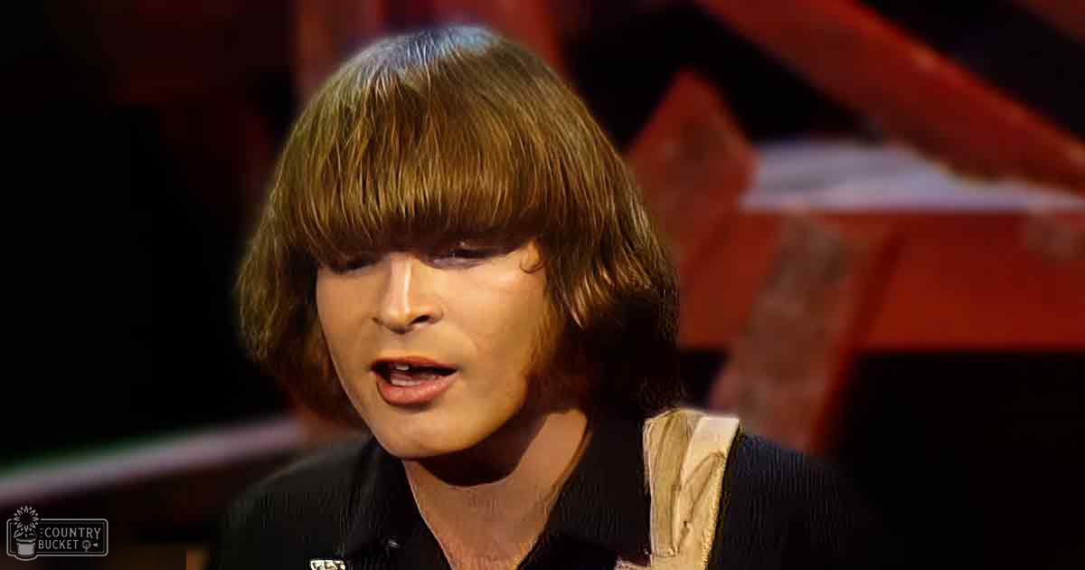A young adult male wearing a dark-colored shirt with a collar, standing indoors near a red piano under warm evening lighting.