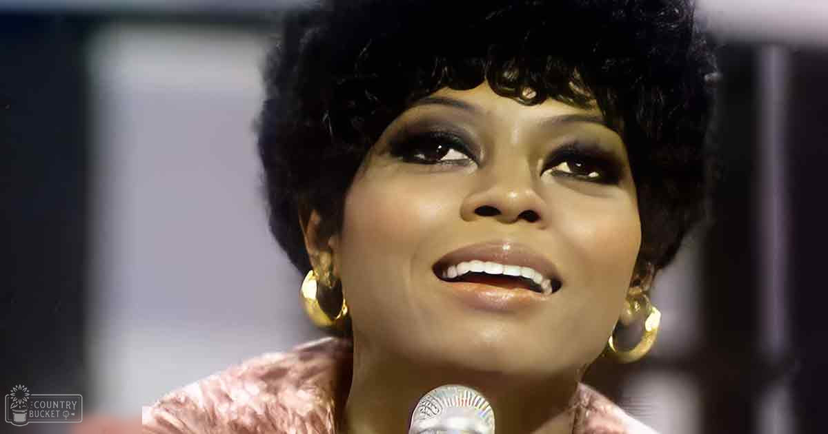 An adult female singer wearing a textured pink blouse with ruffled shoulders, long sleeves gathered at cuffs, large hoop earrings, singing into a microphone on stage.