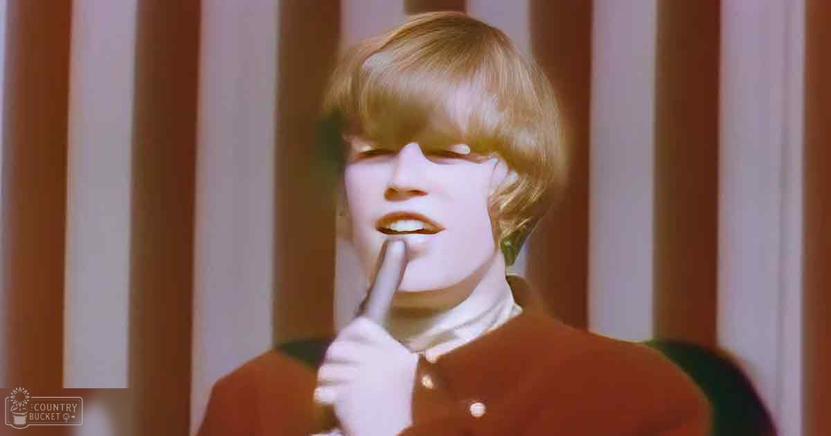 A young adult with short light-colored hair wears a white high-collared shirt under an orange jacket with a star emblem, suggesting a retro style from the 1960s or 70s. Indoors with vertical blinds and natural light.