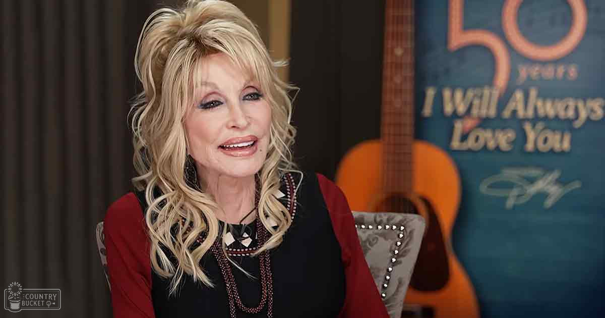 A middle-aged woman in a black top with intricate patterns, paired with a red cardigan or jacket, seated indoors with decorative furniture and a guitar in the background.