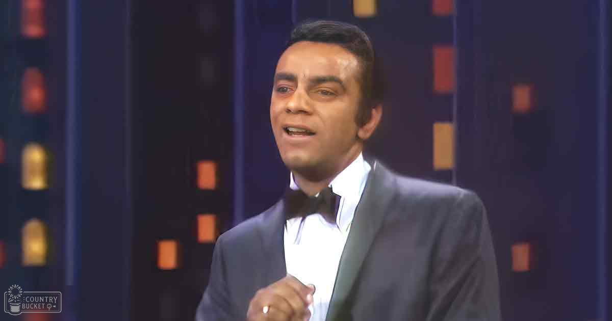 A formal adult male in a tailored, dark gray suit with a white shirt and bow tie, standing against a backdrop of vertical rectangular shapes suggesting an indoor stage or studio environment. The setting includes a dark blue background with orange elements.