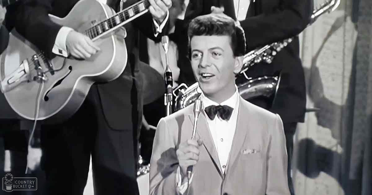 A young adult male singer in a mid-20th-century light-colored suit jacket with a white shirt and black bow tie performs on stage, accompanied by two other band members dressed similarly. The setting is an indoor performance venue with bright lighting focused on the performer.