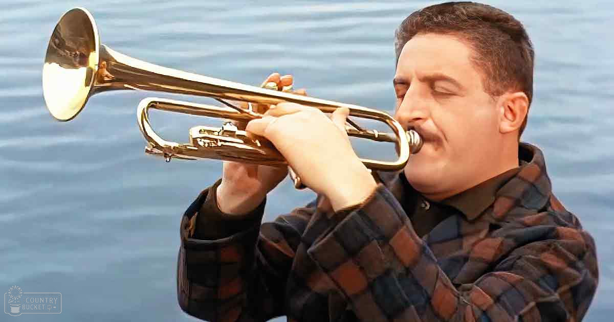 A middle-aged adult male wearing a plaid jacket plays the trumpet near water at what appears to be either early morning or late afternoon, with soft lighting and blue tones dominating the scene.