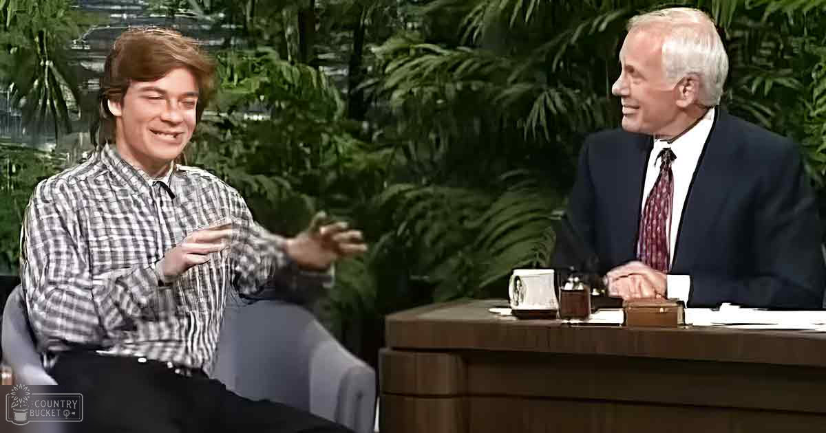 A young adult male in a plaid button-up shirt gestures while seated at a wooden desk with an older adult male wearing a dark suit jacket and patterned tie, engaging in conversation. The setting is a talk show environment with greenery in the background, suggesting an indoor garden-style backdrop.