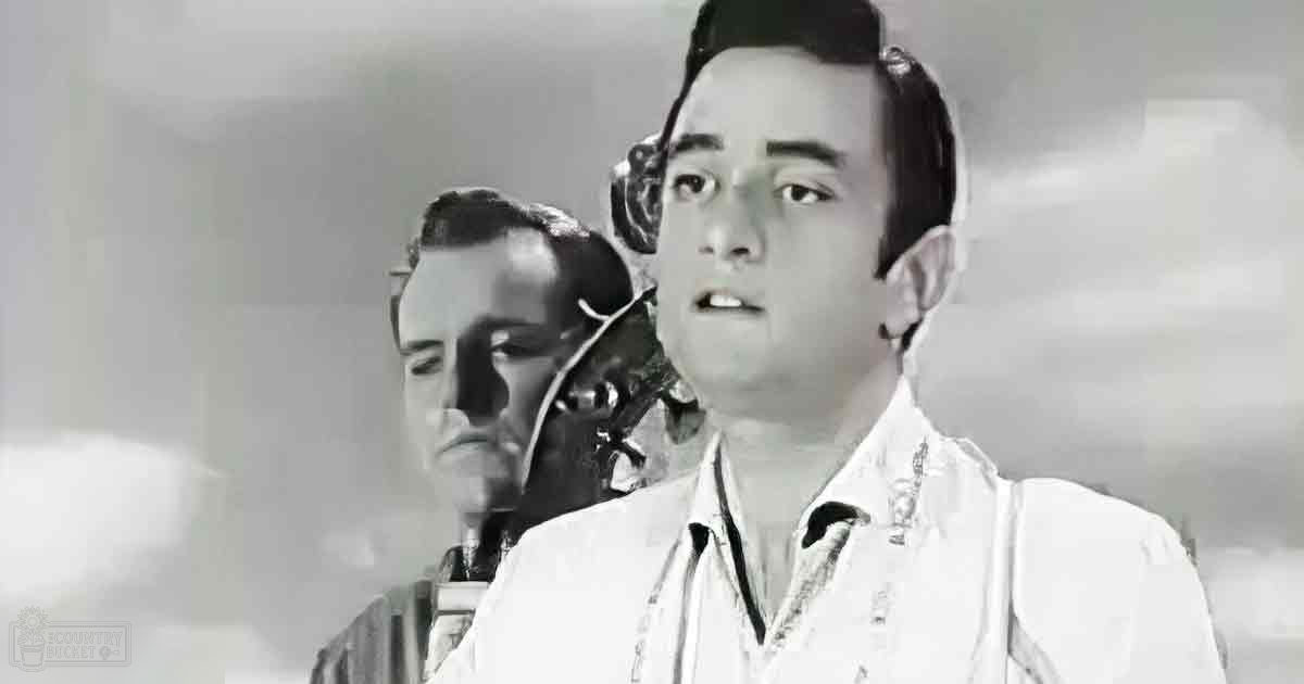 A young adult male wearing a patterned white shirt stands indoors with an adult male in the background; the setting appears to be a room or studio, possibly during daytime with natural light.