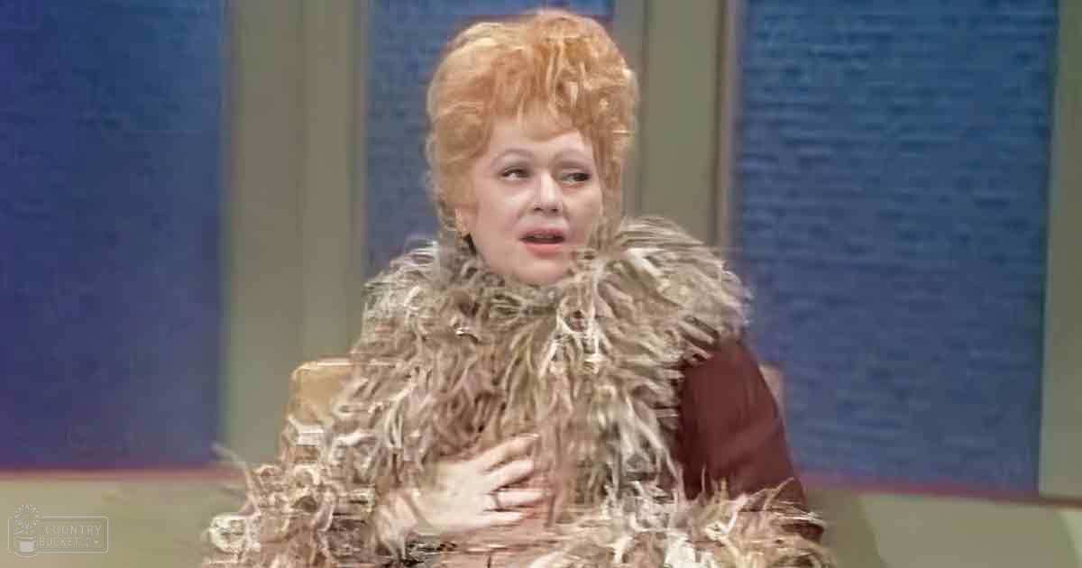 A middle-aged woman stands indoors, wearing a brown fur-like garment with ruffled details around the shoulders against a blue backdrop.