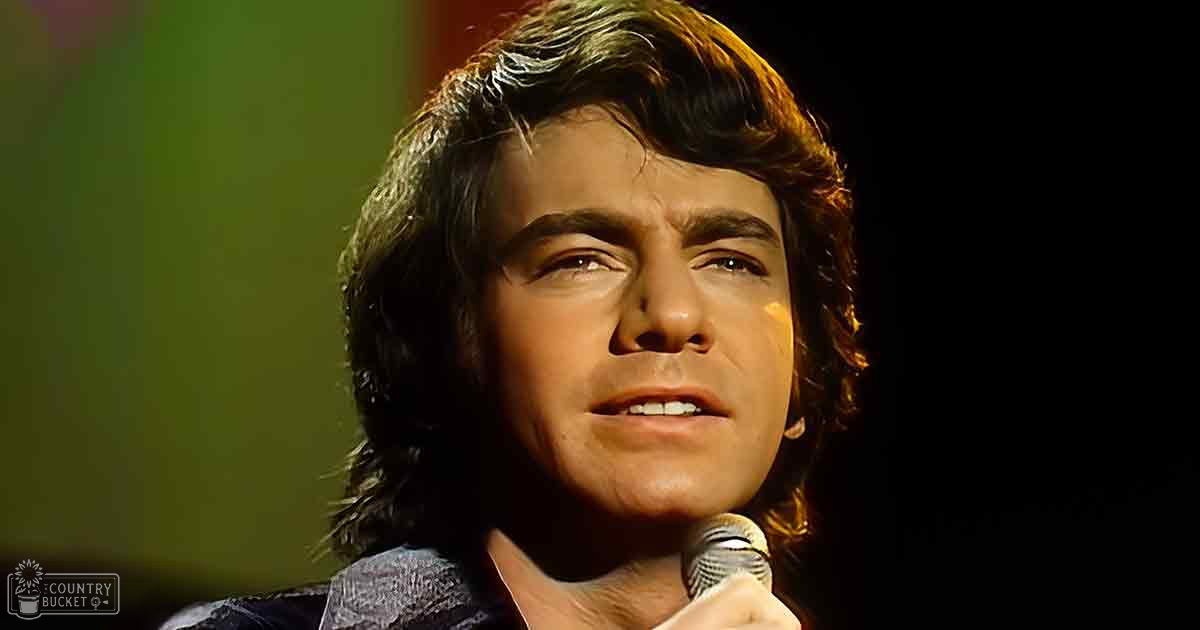 A young adult male singer performing on a dimly lit indoor stage with dark tones dominating the scene.