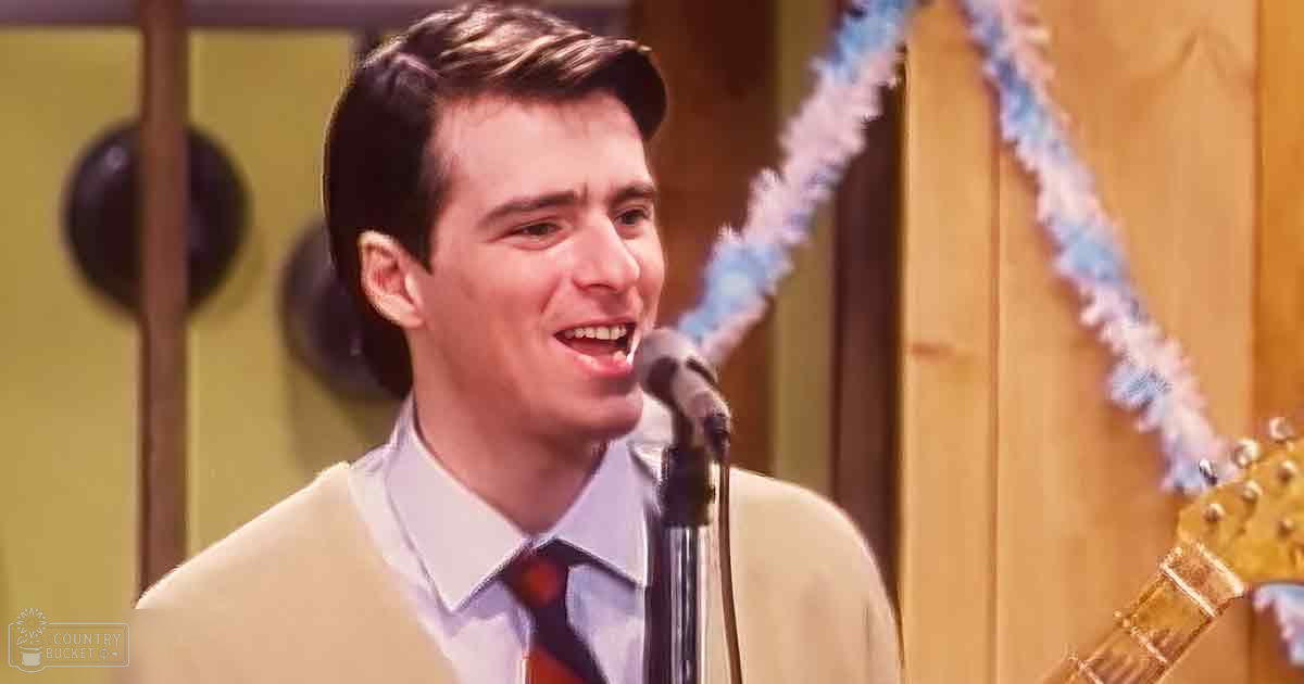 A young adult male in a relaxed fit sweater over a collared shirt with a tie, standing indoors near a microphone stand and guitar, suggesting a performance or rehearsal space.