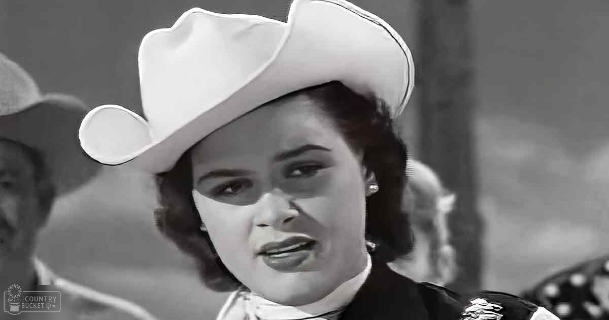A young adult female wearing a white cowboy hat with a wide brim, standing indoors in neutral surroundings under natural daylight.