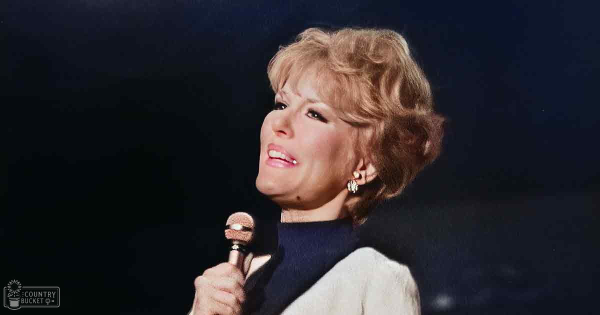 Middle-aged female performer holding a microphone on an indoor stage, wearing a dark blue turtleneck sweater with a fitted cut and a light-colored cardigan from the mid-20th century.