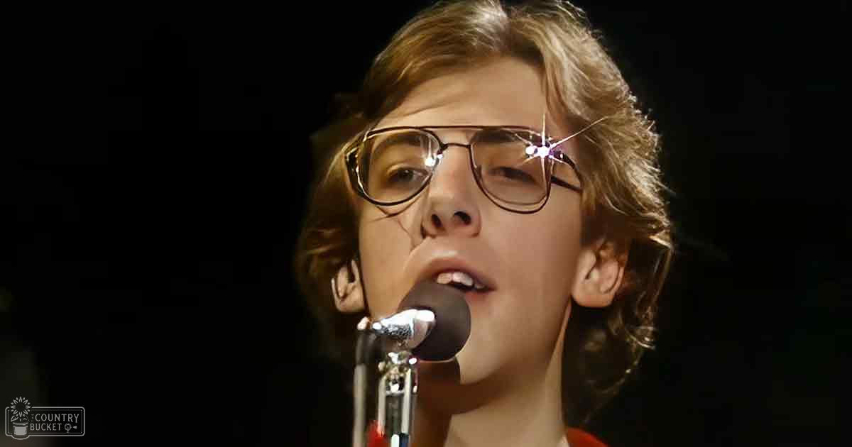 A young adult male wearing a bright red, collared shirt with thin black-framed glasses sings into a microphone at an indoor event.