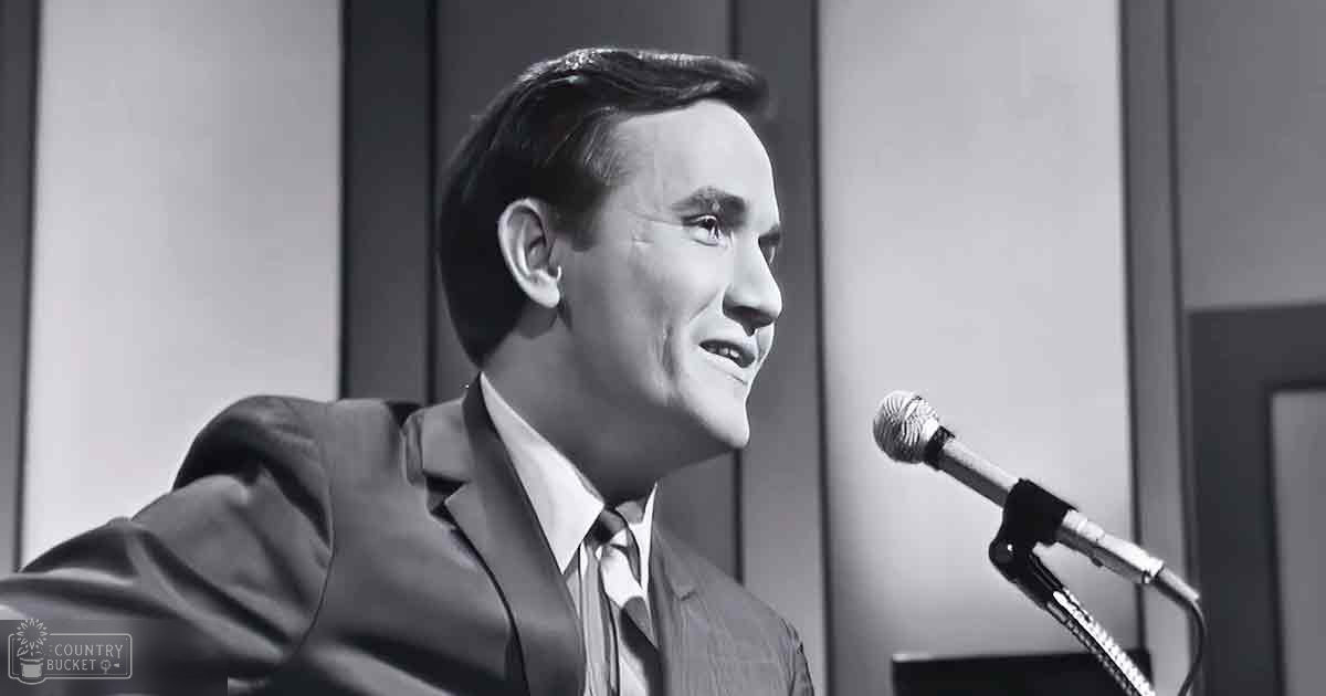 An adult male in a suit jacket and tie, standing indoors with a microphone to his right.