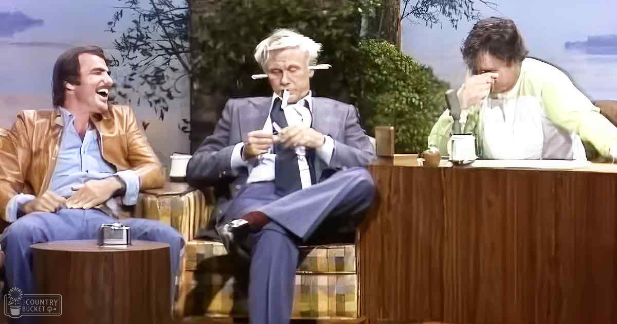 Adult male in shiny gold jacket on talk show set with older male in gray suit smoking, middle-aged female in light green cardigan seated at desk; indoor studio designed to look like outdoor scene with trees and water, suggesting spring or summer day.