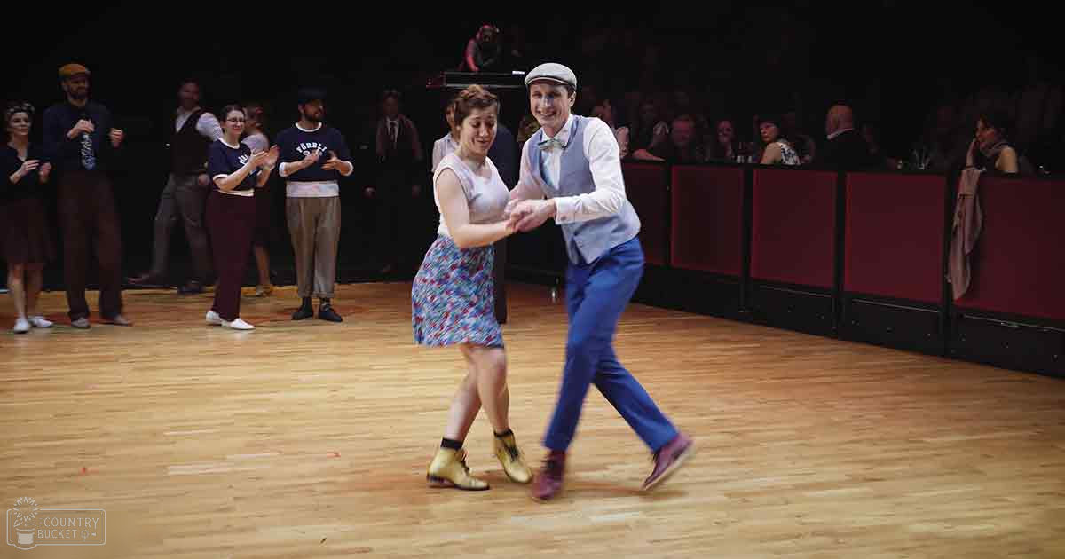 A young adult male and female couple dancing in an indoor dance hall or theater. The man wears a light blue vest over a white shirt with a bow tie, dark blue pants, red shoes, and a white cap. The woman sports a sleeveless dress with shades of blue and pink floral patterns, yellow boots, and her hair tied back.