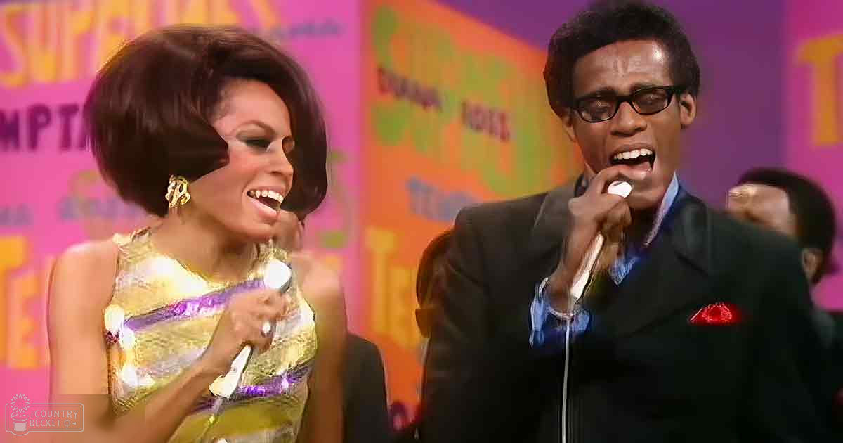 An adult female singer wearing a shimmering gold dress with purple accents performs on stage alongside an adult male musician dressed in a dark suit with a red pocket square, both holding microphones against a backdrop of colorful banners indicating a staged event or performance.