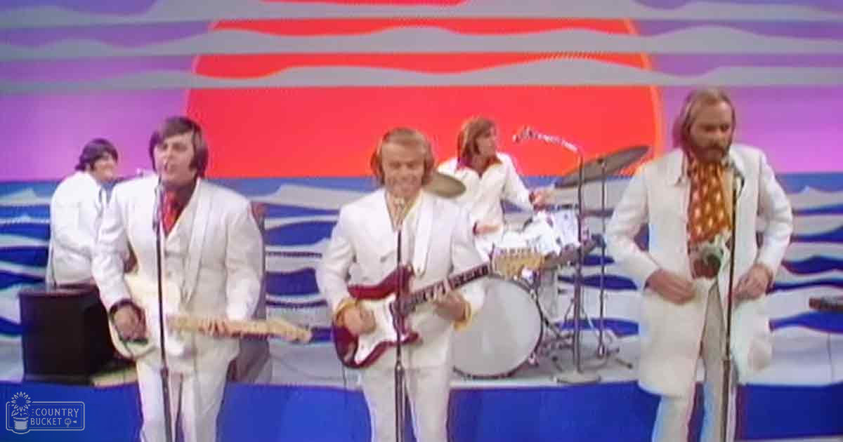 Musical performance group in 1970s indoor stage setting with colorful backdrop, bright lighting, and performers wearing white suits.