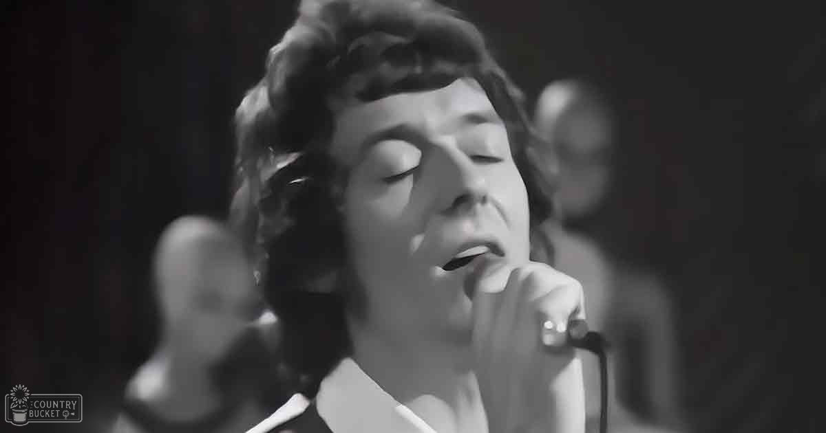 A female adult singer wearing a white, high-collared garment with rings on her fingers sings into a microphone in an indoor performance venue under dim stage lighting.
