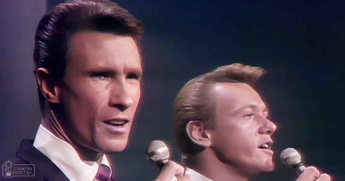 An adult male singer performing on stage with a patterned tie, accompanied by another adult male performer wearing a light-colored suit jacket.