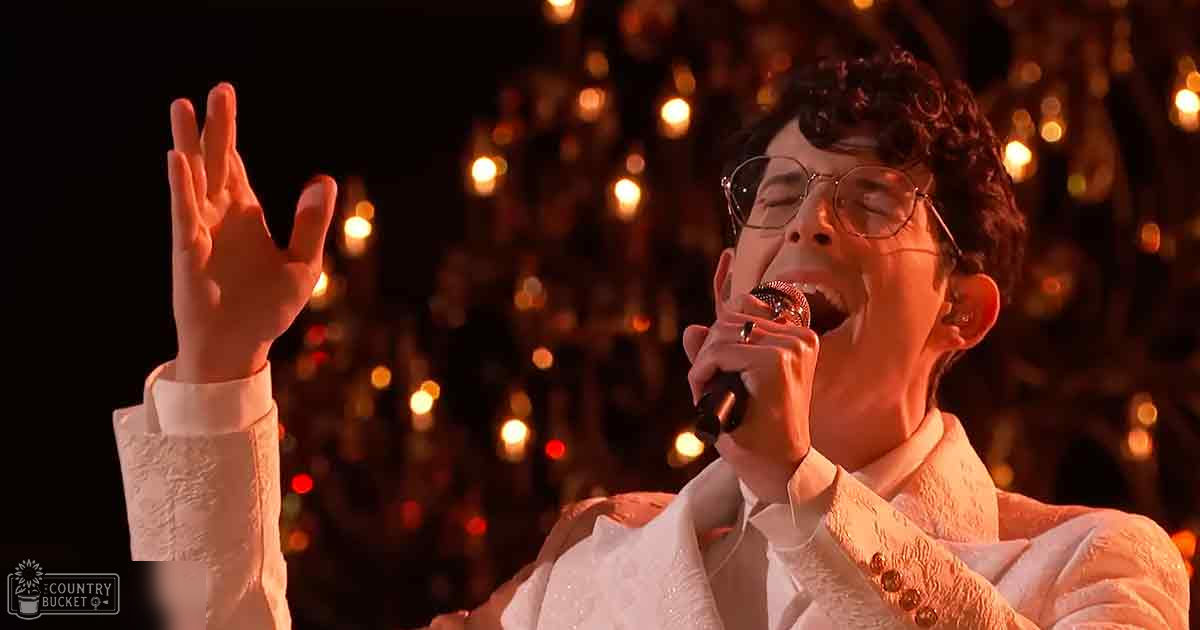 A young adult male performer wearing a white tailored suit jacket and shirt, holding a microphone on stage under stage lighting.