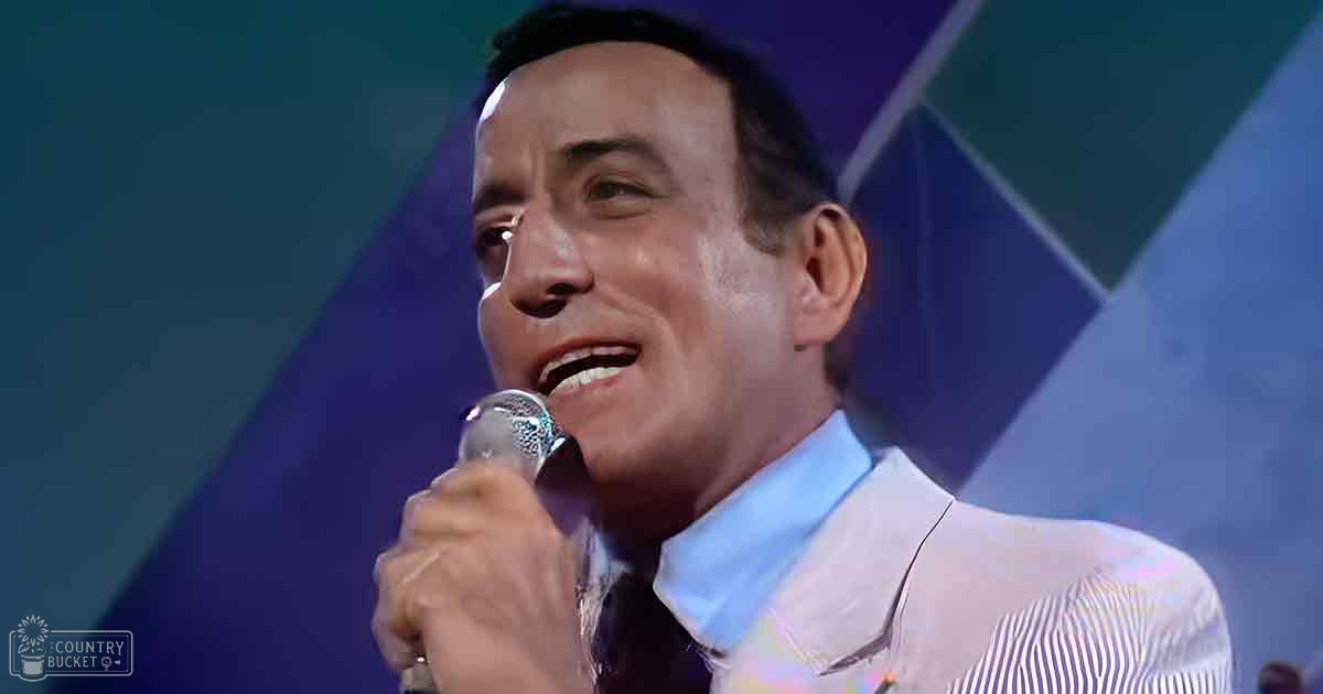 A middle-aged male singer performing, wearing a light-colored striped shirt with cotton-like fabric, holding a metallic microphone against an abstract blue and green geometric backdrop in what appears to be an indoor stage setting.
