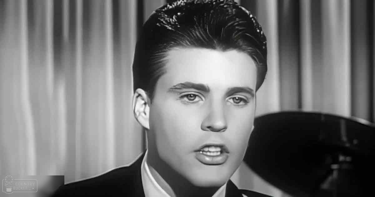 A young adult male in a dark suit jacket stands before a piano, wearing a white shirt, in an indoor setting with curtains and neutral tones.