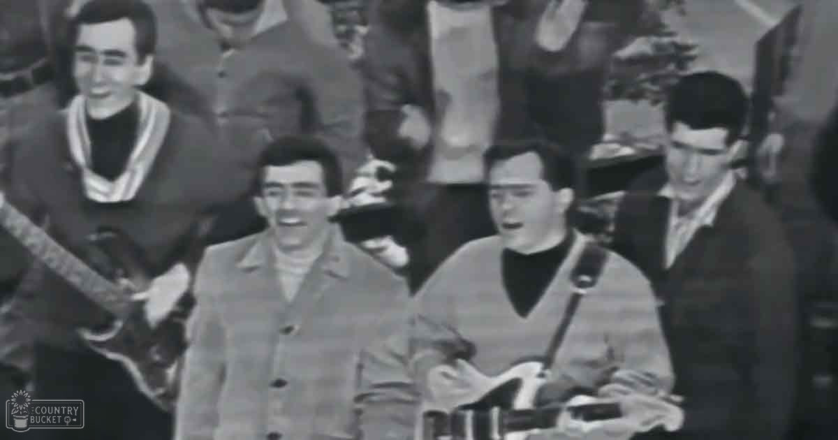 A musical performance in an indoor setting, with young adult males wearing dark jackets with light-colored trim around collars and shoulders. The background shows a crowd of spectators under natural or ambient indoor lighting.
