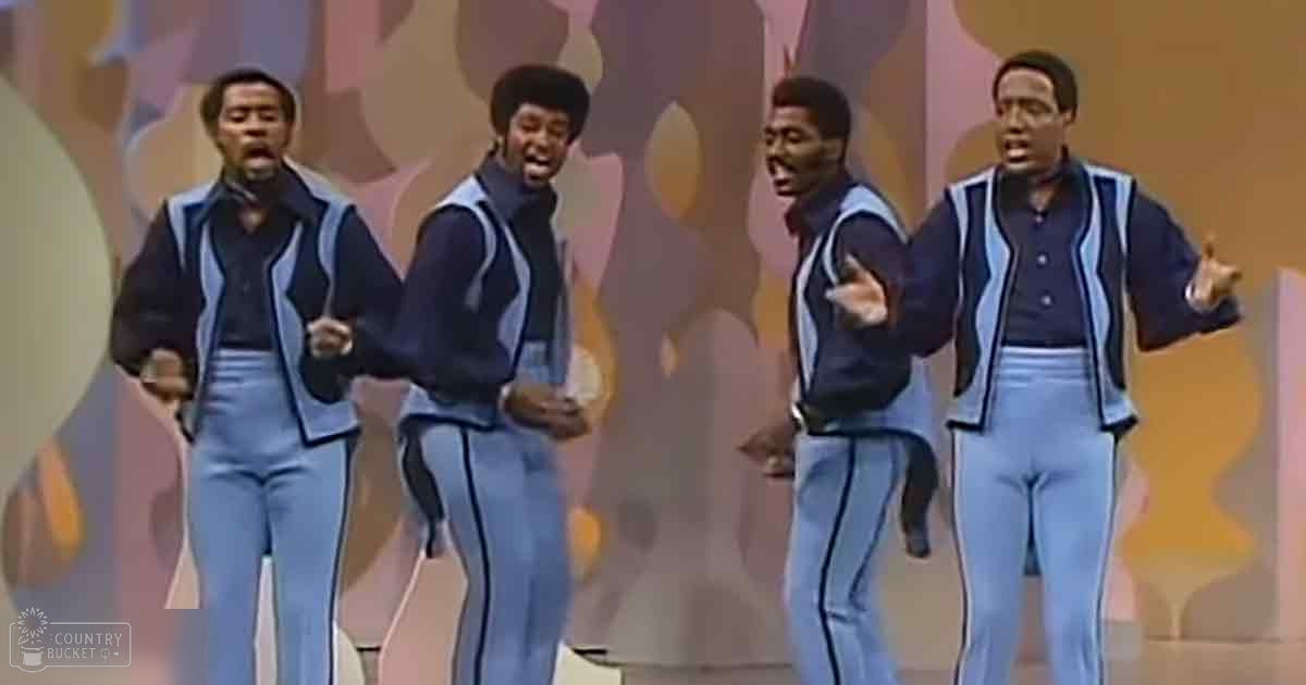 Young adult male performers wearing 1970s-style light blue jumpsuits with dark blue stripes, performing on a stage with colorful abstract backdrop.