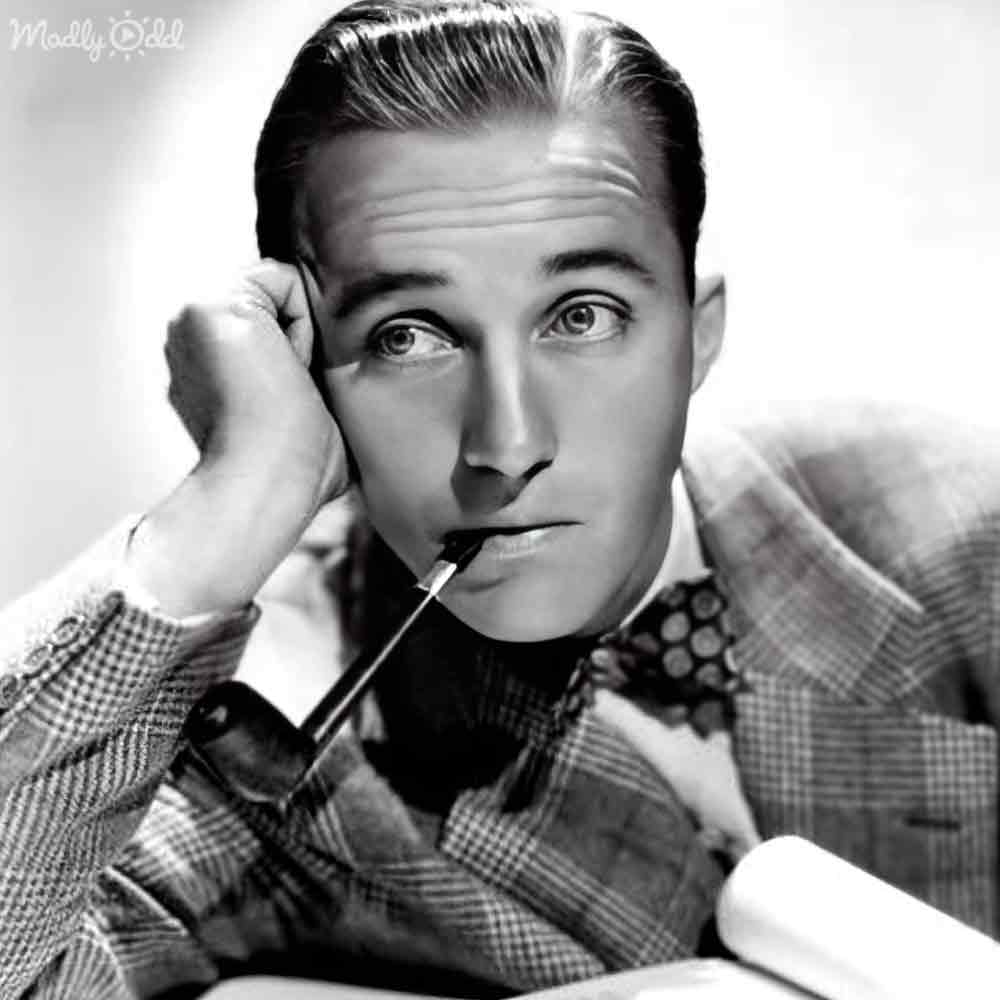 A mid-20th century adult male in a checkered blazer and collared shirt, holding a cigarette holder with one hand resting on his face against a plain white backdrop.