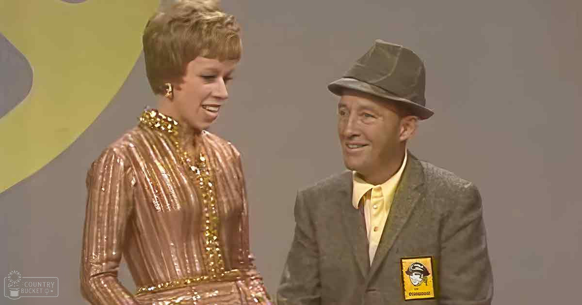 A comedic duo in a studio setting: one person wears a shiny, sequined dress with vertical stripes and gold embellishments; the other dons a gray blazer over a yellow shirt with 'Sailor' text and a sailor's hat patch.