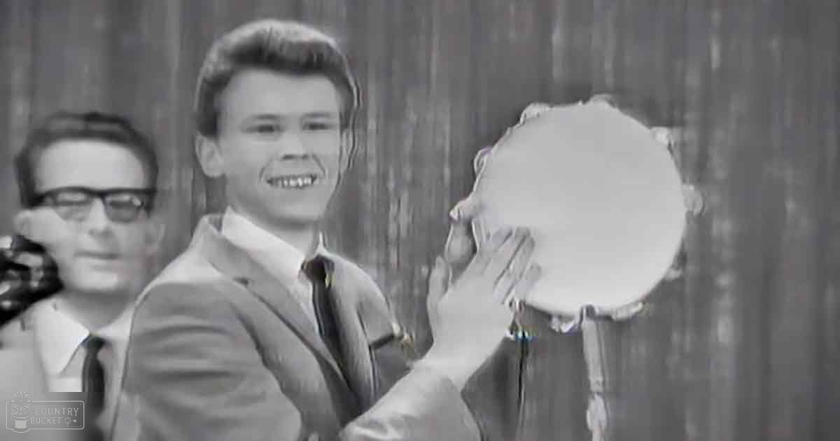 Young adult male in light-colored suit jacket and tie, mid-20th century style, with a microphone in an indoor studio setting.