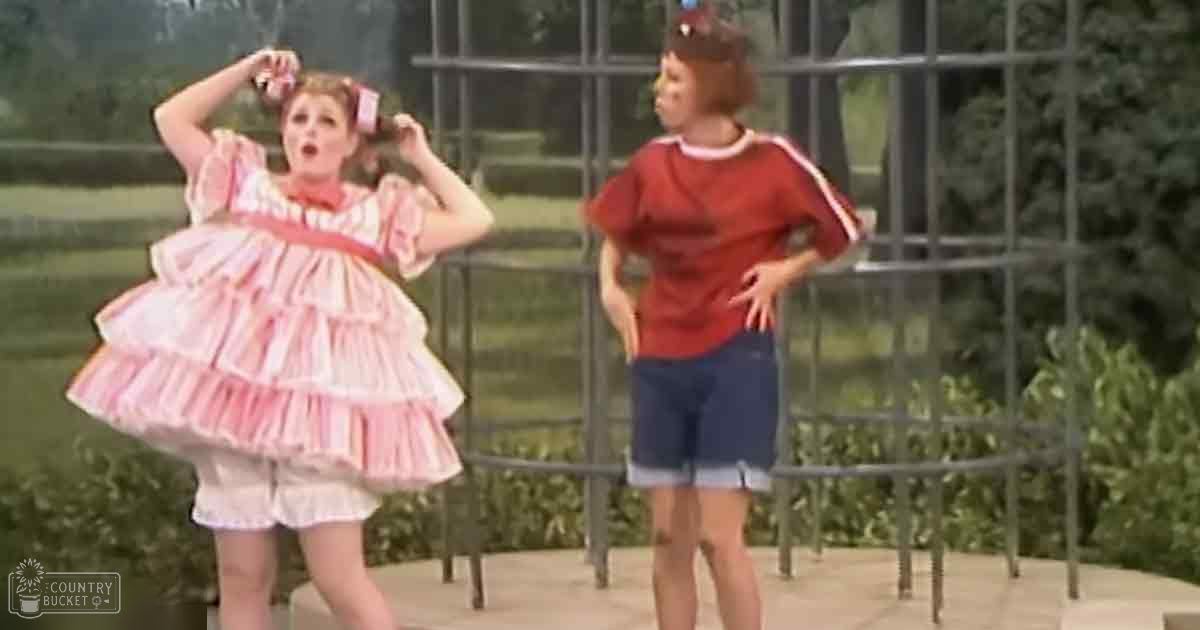 A child dressed as a princess and another child wearing a casual outfit stand outdoors with an arbor-like structure behind them, surrounded by greenery under bright daylight.