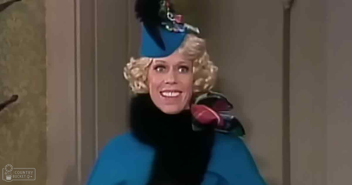 An adult female in a tailored blue coat with black fur trim and red high-heeled shoes stands indoors against neutral-colored walls, suggesting a residential or professional environment.