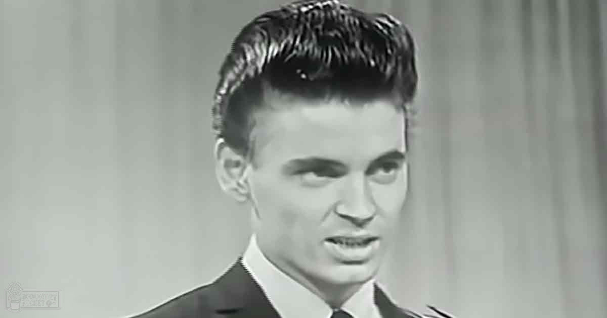 A young adult male wearing a dark-colored suit jacket and white shirt in a black-and-white indoor setting resembling a studio with neutral lighting.