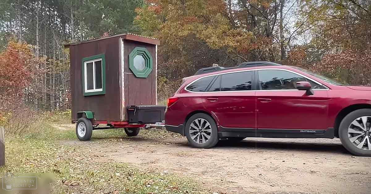 A small brown trailer with green trim, towed by a red Subaru Outback on an outdoor dirt path surrounded by trees with fall foliage.