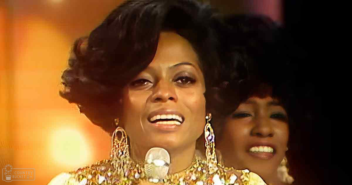 An adult female singer in a form-fitting, intricately detailed garment with large ornate earrings, standing on an indoor stage with warm lighting and formal attire.