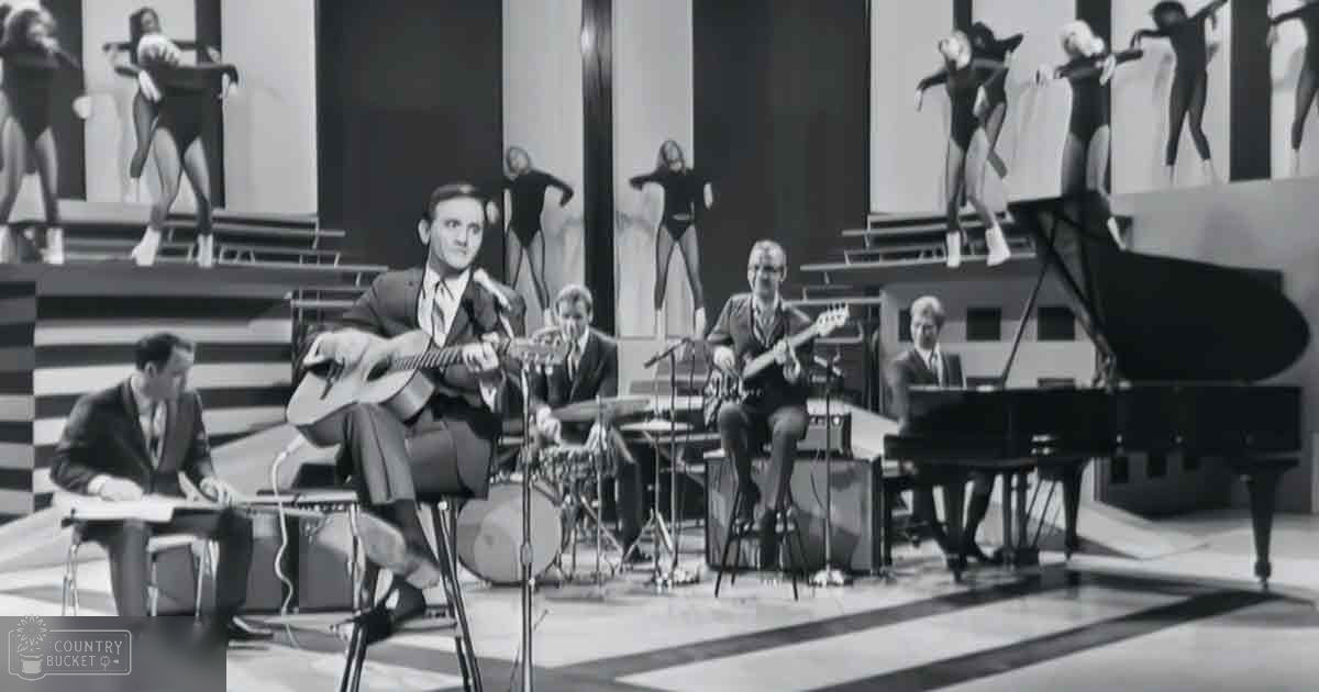 A mid-20th-century musical performance with a young adult male singer in the foreground and other musicians playing instruments on stage under warm indoor lighting.