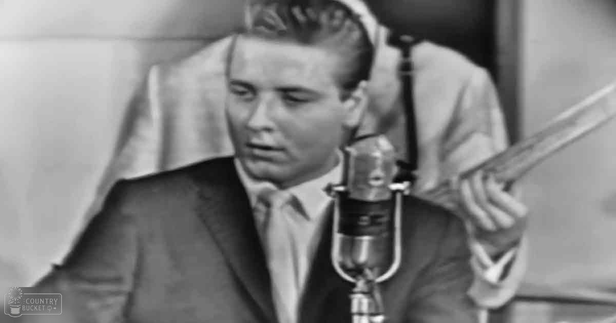 A young adult male wearing a tailored dark suit jacket, light-colored shirt with a tie, holding a guitar in an indoor recording studio environment.