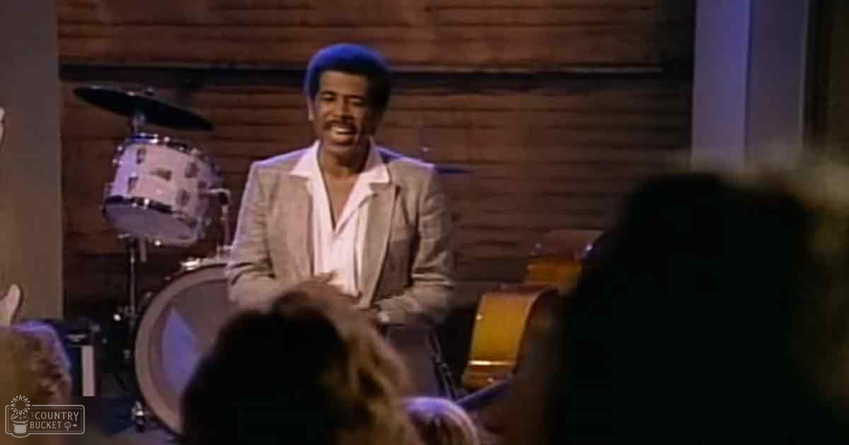 A middle-aged male singer in a light-colored suit jacket performing at what appears to be a music venue with wooden paneling, stage lighting focused on the performer.