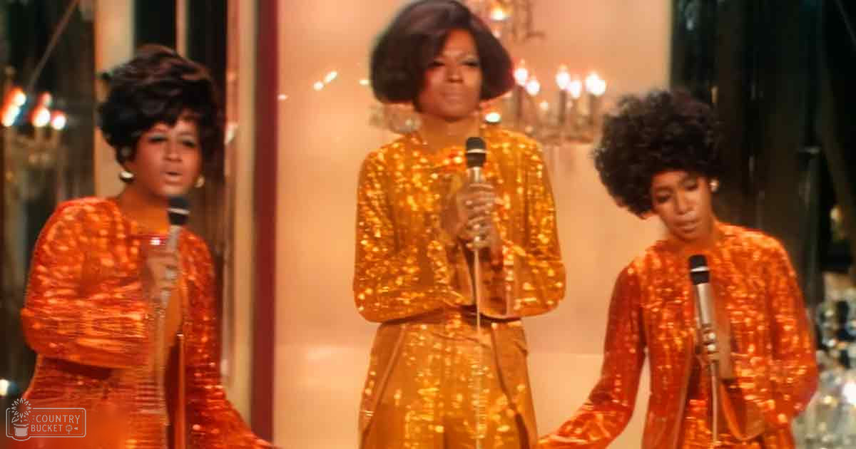 Three adult females performing on stage, dressed in matching 1960s-style sequined orange dresses, with ornate lighting and microphones indicating a formal event space.